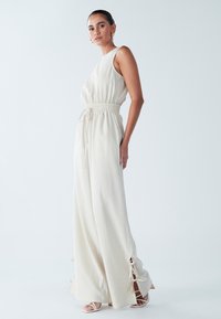 Kremasto-bež kombinezon z vrvico v pasu, širokimi hlačnicami, stranskimi razporki in subtilno teksturo. Kombiniran z belimi sandali na visoki peti.