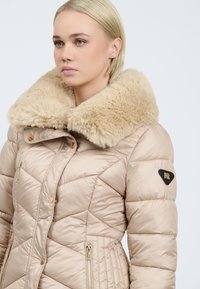 Beige polstret jakke med en stor faux fur krave, skråt quiltet design, guld hardware detaljer og sidelommer med lynlås.