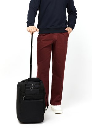 Black wheeled backpack with multiple zipped pockets, soft fabric, and a retractable handle, alongside a person in burgundy pants and white shoes.