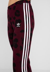 Bordowe legginsy w czarny lampartowy wzór, z trzema pionowymi białymi paskami i wyszywanym czarnym logo Adidas po boku.