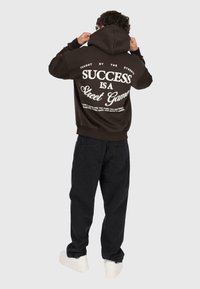 Personne de dos portant un hoodie noir avec le texte "Le succès est un jeu de rue" et un pantalon noir, ajustant la capuche avec les deux mains.