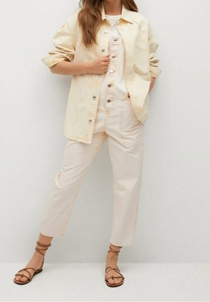 Veste en denim jaune clair portée sur un t-shirt blanc avec une salopette beige ; le pantalon présente des coutures claires. Des sandales à lanières marron clair complètent le look.