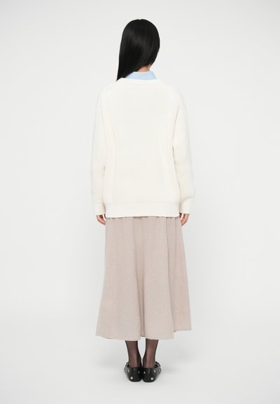 Camisola leve de malha cremosa com textura canelada, combinada com uma saia fluida bege. Sapatos pretos com detalhes prateados completam o visual.