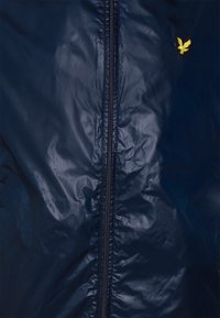 Navy blue, shiny jacket with a full zip closure. Features a small yellow eagle logo on the left side. Smooth texture with no visible patterns.