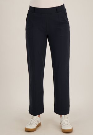 Marineblauwe broek met rechte pijpen en voorzakken gedragen met witte sneakers bij een persoon tegen een effen achtergrond.