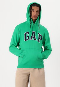 Felpa verde con logo "GAP" in blu navy. Presenta una tasca frontale e cappuccio con cordino. Realizzata in materiale morbido con maniche lunghe.