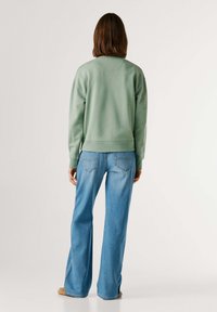 Πράσινο φούτερ με μακριά μανίκια και ριμπ μανσέτες, σε συνδυασμό με ανοιχτό μπλε τζιν με φαρδιά παντελόνια, απεικονιζόμενο από πίσω σε απλό φόντο.