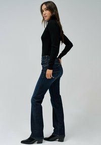 Blusa negra de manga larga combinada con jeans de campana oscuros. Los jeans tienen costuras visibles y bolsillos, y un ligero desvanecimiento en las rodillas. Botas negras de tacón.