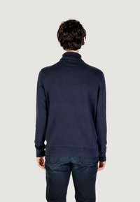 Maglione lavorato a maglia blu navy con colletto alto, maniche lunghe e polsini a coste; indossato con jeans scuri, visto da dietro.