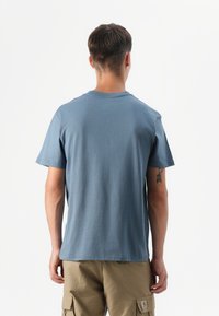 Ung mann som står med ryggen til, iført blå t-skjorte og khaki cargo-shortser mot en enkel hvit bakgrunn. Synlig tatovering på venstre arm.