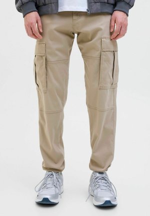 Mann trägt beige Cargohosen mit Seitentaschen, graue Jacke, weißes Hemd und weiße Sneaker und steht vor weißem Hintergrund.