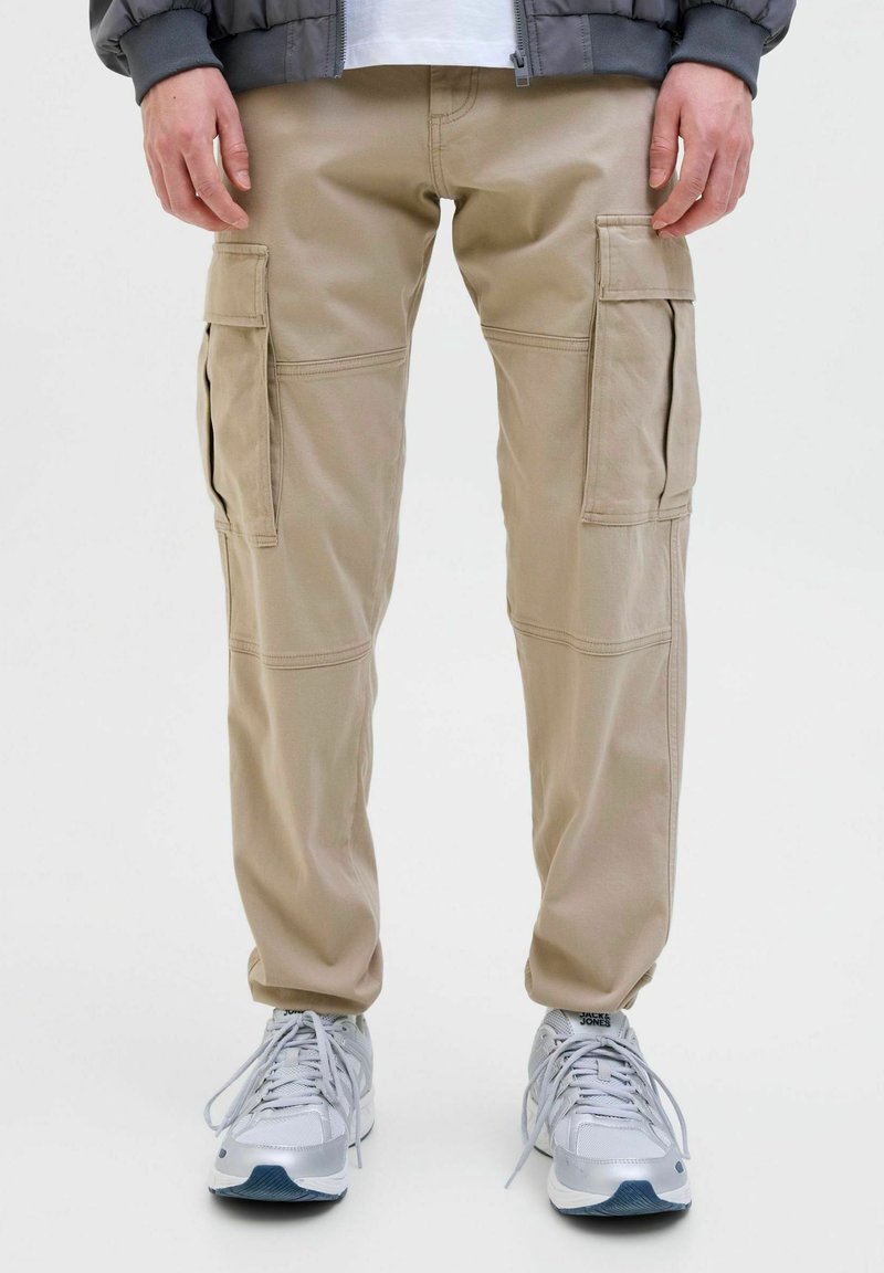 Homme portant un pantalon cargo beige avec des poches latérales, une veste grise, une chemise blanche et des baskets blanches, debout sur fond blanc.