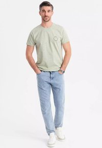 Camiseta verde claro de manga corta con un patrón moteado y un logo redondo, combinada con jeans de mezclilla azul claro y zapatillas blancas.