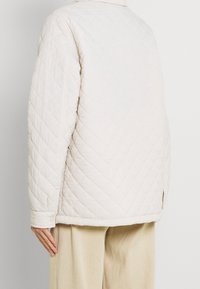 Beżowa pikowana kurtka o luźnym kroju, z przeszyciami w kształcie diamentów, rozporkami po bokach i ozdobnymi guzikami na mankietach.