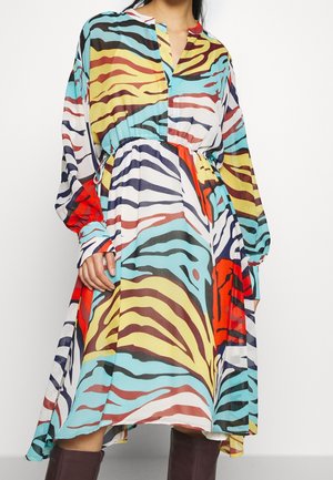 Robe à manches longues avec des rayures zébrées abstraites colorées en rouge, jaune, bleu, blanc et noir, taille cintrée, portée avec de longues bottes marron.