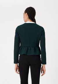 Dark green peplum jacket with long sleeves, textured fabric, crew neckline, and a ruffled hem, paired with black fitted pants.