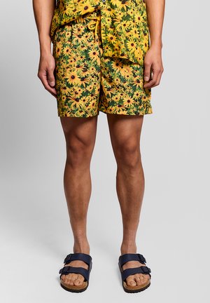 Man die een gele korte broek met bloemenprint en een bijpassend overhemd draagt, met donkerblauwe sandalen met dubbele band, staand tegen een effen achtergrond.