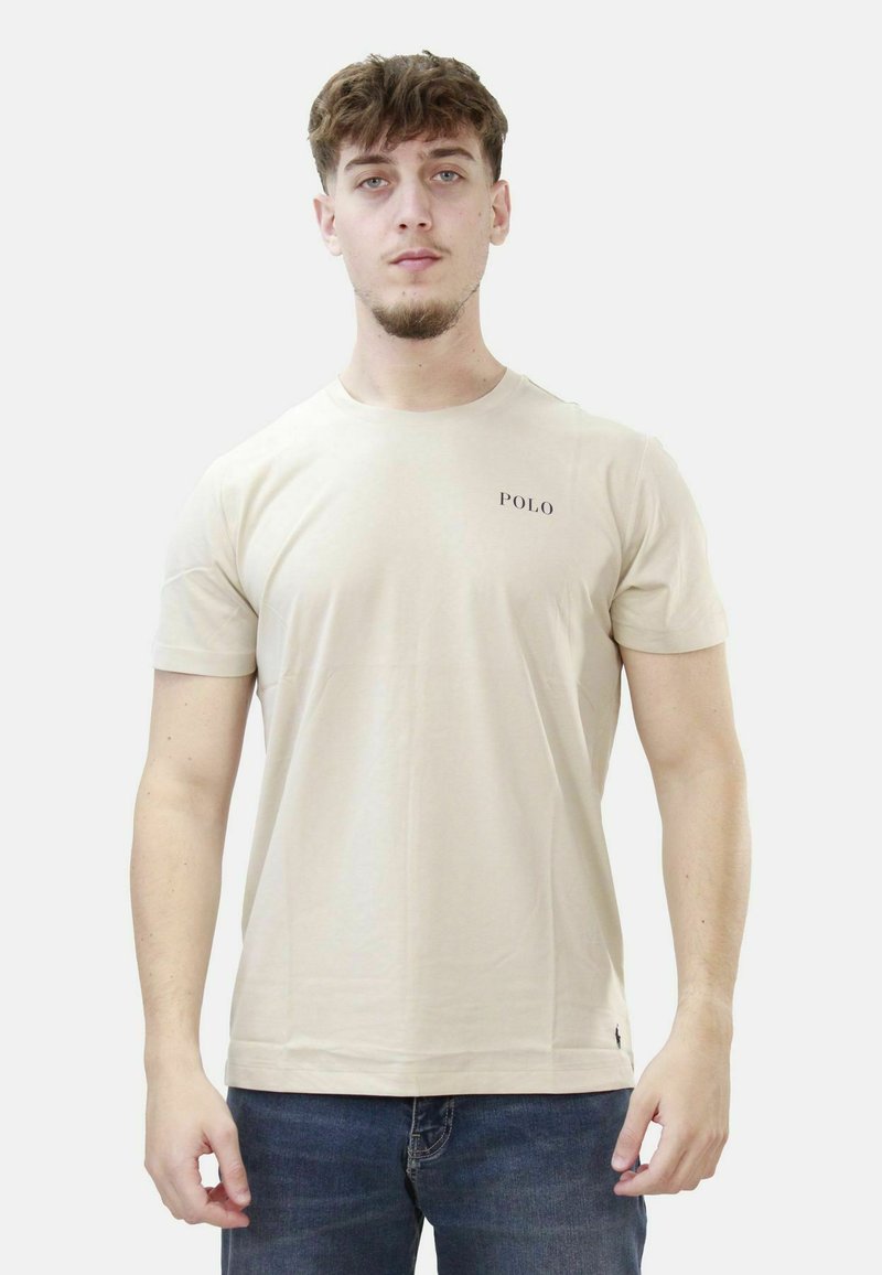 Young man with light brown hair wearing beige Polo t-shirt and dark jeans standing against plain light background.