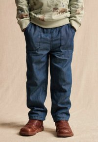 Farkut rentoa mallia, joissa on kaksi suurta etupainiketta ja kapeneva lahje, yhdistettynä ruskeisiin nahkasaappaisiin. Vihreä kuviollinen collegepaita.