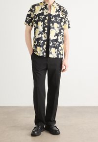 Camisa de botones de manga corta con fondo negro y grandes patrones florales amarillos, combinada con pantalones negros y zapatos negros.