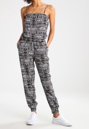 Kobieta w czarno-białym kombinezonie bez rękawów z geometrycznym wzorem, z elastycznymi mankietami i białymi trampkami, z rękami w kieszeniach.
