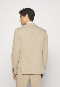 Beige zakó, lekerekített hajtókával, egy hátsó szellőzőnyílással és fekete gombokkal az ujjakon. Simaszerű anyag, testhezálló fazon.