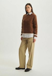Pull en laine brun avec une coupe ample, doté d'une bordure blanche, porté par-dessus une chemise rayée blanche, associé à un pantalon beige et des chaussures noires.