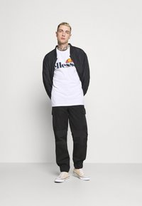 Black lightweight bomber jacket over a white t-shirt with a logo. Black cargo pants with pockets and beige sneakers. Simple, casual outfit.