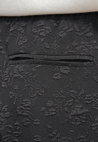 Tissu noir avec broderie florale en relief et une poche fendue horizontale, gros plan montrant les détails de la texture et du motif.