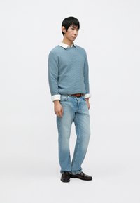 Svetlomodrý textúrovaný sveter cez bielu košeľu s golierom, kombinovaný so svetlomodrými džínsami a tmavohnedými topánkami, stojaci na jednoduchom pozadí.