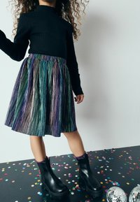 Maglietta nera a coste con maniche lunghe abbinata a una gonna plissettata e brillante multicolore. Stivaletti neri con suola spessa. Coriandoli sul pavimento.