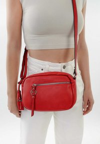 Rød læder crossbody taske med en struktureret finish, lynlåslomme foran, justerbar rem og et mærketag. Målene er omtrent i mellemløsningen.
