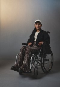 Una persona seduta su una sedia a rotelle, con indosso una giacca nera sopra una maglietta bianca, abbinata a pantaloni a stampa leopardata e un cappellino bianco.