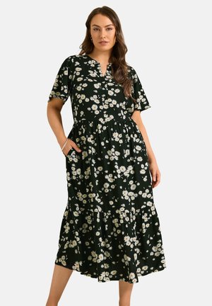 Femme debout les mains dans les poches, portant une robe midi noire à imprimé floral marguerite blanche et manches courtes.