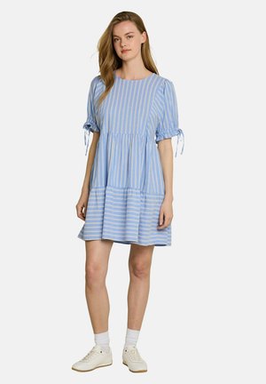 Junge Frau trägt ein blau-weiß gestreiftes kurzes Kleid mit Puffärmeln, die am Ellbogen gebunden sind, weiße Socken und weiße Turnschuhe.