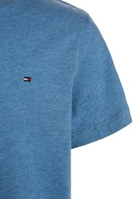 Niebieska bawełniana koszulka z krótkim rękawem, z małym haftowanym logo w kolorach czerwonym, białym i czarnym po lewej stronie w pobliżu klatki piersiowej.