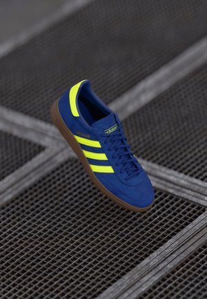 Baskets Adidas bleues avec des rayures jaunes et une semelle marron flottant au-dessus de grilles métalliques.