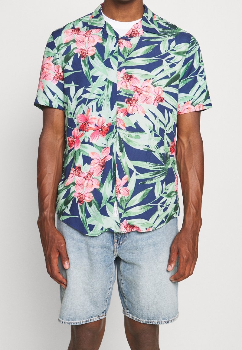 Kurzärmeliges Hemd in Marineblau mit großem pinkfarbenem Blumen- und grünem Blättermuster; Knopfleiste; kombiniert mit hellen Denim-Shorts.