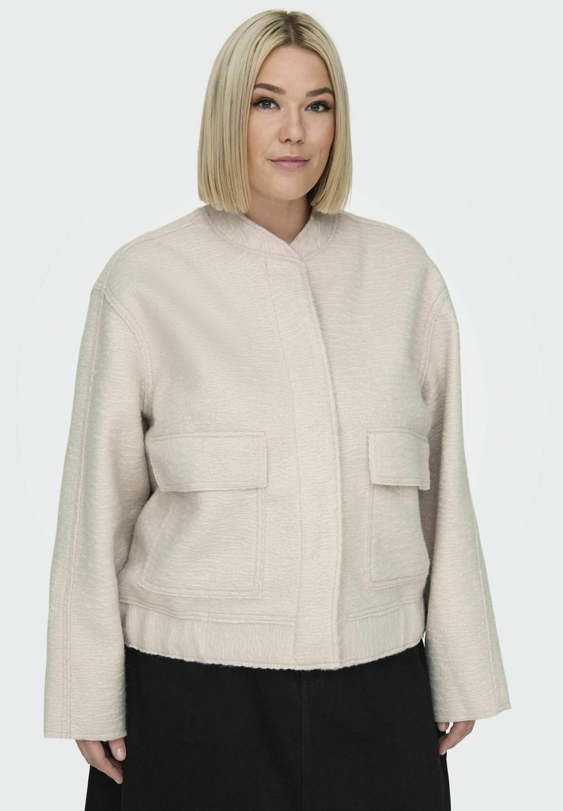 Ljus beige, kort jacka med ett texturerat tyg, ståkrage och två framfickor. Ärmarna är vida och designen är boxig.