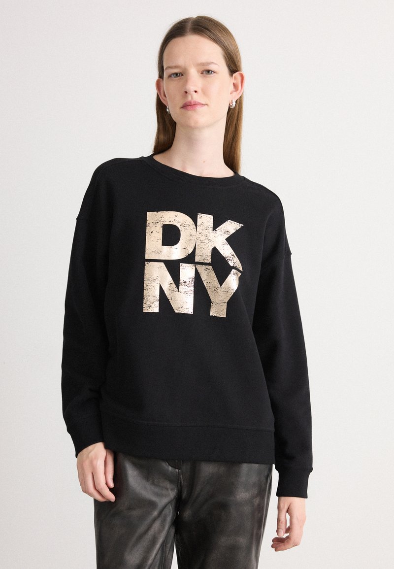 Svart hettegenser med overdimensjonert, slitt gull "DK NY"-logo. Lange ermer, rund hals, avslappet passform, kombinert med glinsende mørke bukser.