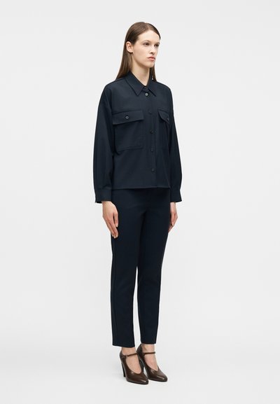 Γυναίκα που φοράει μια υπερμεγέθη μπλε σκούρα πουκαμίσα με τσέπες με καπάκι και εφαρμοστά παντελόνια στο ίδιο χρώμα, συνδυασμένα με σκούρα καφέ πλατφόρμες Mary Jane.