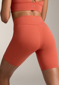 Piros-narancs magas derékú biciklis sort rugalmas, sima anyagból, széles derékrésszel és térd közepéig érő дизайнер. Diszkrét márkalogó látható.