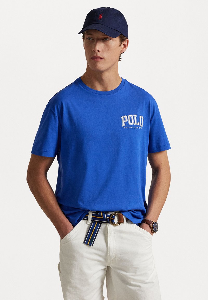 Kungsblå bomull t-shirt med "POLO"-logotyp och vita accenter, i kombination med krämfärgade byxor och ett randigt bälte. Mörk keps kompletterar outfiten.