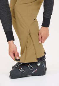 Tan skibroeken met ritsen bij de enkels, gecombineerd met zwarte skischandchoenen met verstelbare gespen en een textuurdesign.
