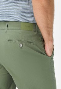 Pantaloni di cotone verdi con una texture liscia, dotati di una tasca posteriore con un bottone argentato e una patch in gomma color oliva.