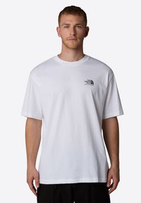 Giovane uomo in piedi di fronte, che indossa una maglietta bianca a maniche corte con un piccolo logo nero "The North Face" sul lato sinistro del petto.