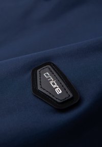 Étiquette en tissu noire cousue avec le mot "onbre" en blanc sur un fond textile bleu foncé et lisse.