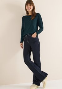 Dlouhý rukáv s vytisknutým vzorem v barvě modrozelené, spárovaný s tmavě modrými zvonovými džínami. Džíny mají světlé švy a jsou nošeny s krémovými teniskami.