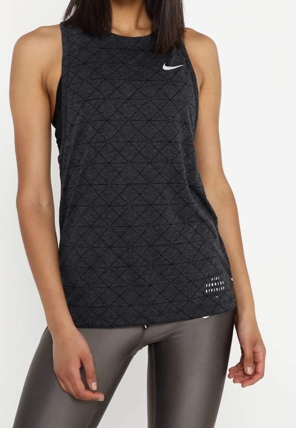 Femme portant un débardeur noir sans manches Nike avec un motif géométrique et des leggings gris, debout sur un fond blanc.