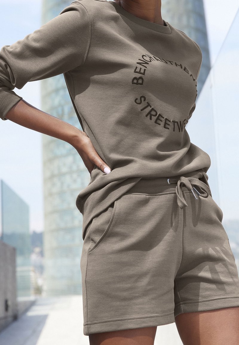 Femme portant un sweat-shirt beige et un short assorti avec un texte circulaire, debout à l'extérieur avec des bâtiments urbains en arrière-plan.